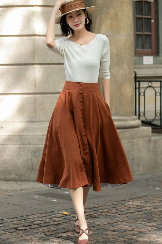 A woman wearing a button down midi linen skirt in brown with a white top, paired with a brown hat and standing on a cobblestone street.