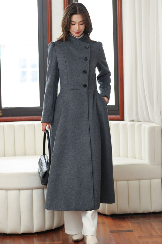 A woman standing in a room with a window, wearing a dark grey A-line wool coat with long sleeves and a button closure, paired with white pants.