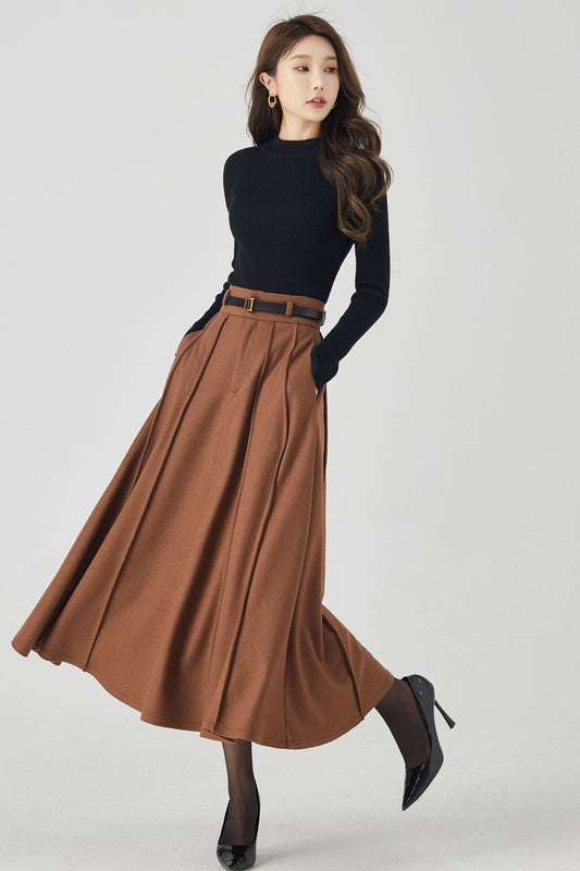 A woman modeling a brown pleated high-waisted wool midi skirt with a black top and dark shoes, against a plain background.