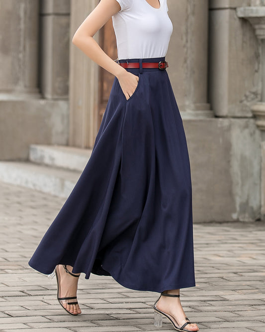 A woman wearing a blue A-line long linen skirt with side pockets, paired with a white top and a brown belt, standing on a cobblestone street.