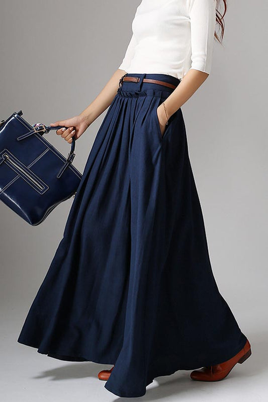 A navy blue linen maxi skirt with a high-waisted design, pleated waist, and side pockets. The skirt is ankle-length and the outfit is accessorized with a belt and handbag.