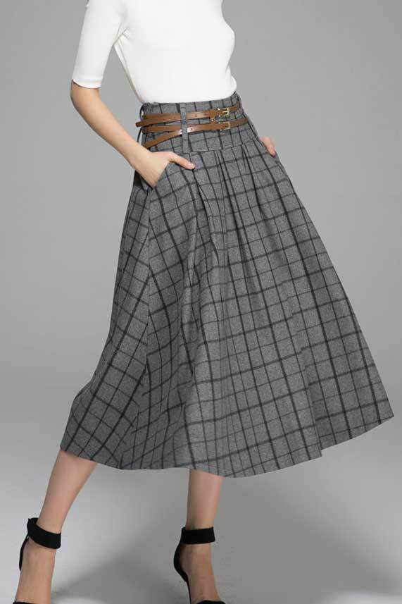 A woman wearing a dark gray pleated midi skirt with a wide waistband and twin side pockets, paired with a black top and black shoes. Accessory belt with a brown buckle is visible.