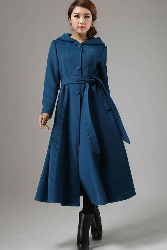 A woman standing wearing a blue wool swing coat with a hood, long sleeves, and a belt waist.