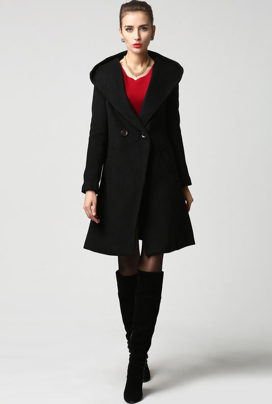 A woman standing and modeling a black wool coat with a big hood, two big pockets, and button fastening through the front. She is also wearing a red top and tall boots.
