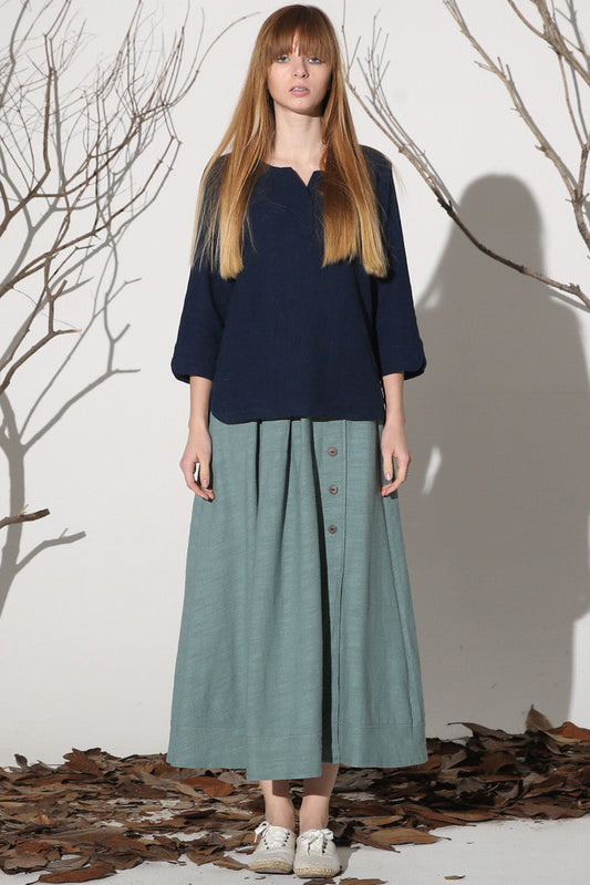 A woman standing in a natural setting wearing a maxi linen skirt with buttons on the front and pockets on the sides. The skirt is green and she is paired with a navy top.