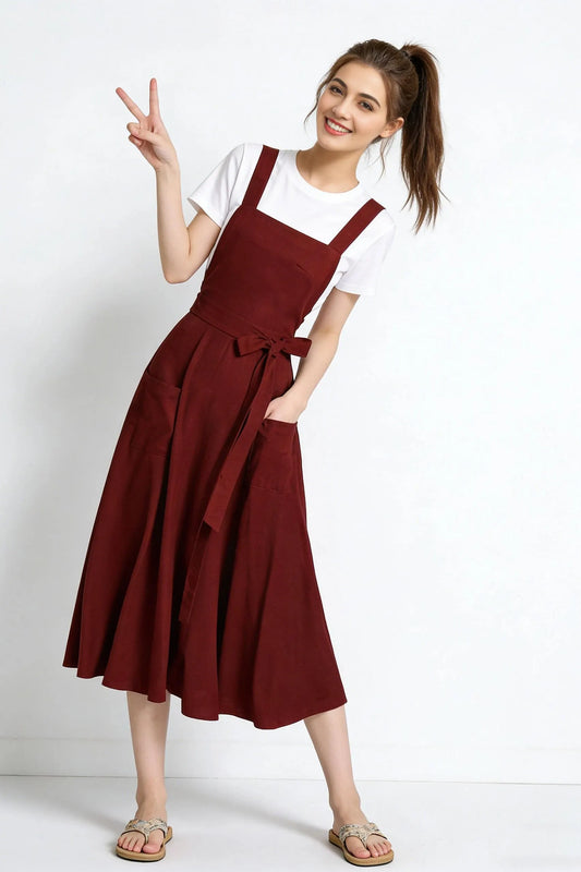 May include: A burgundy-colored overall dress with a white t-shirt underneath. The dress features a square neckline, wide straps, a waist tie, and large pockets. The model is wearing sandals.