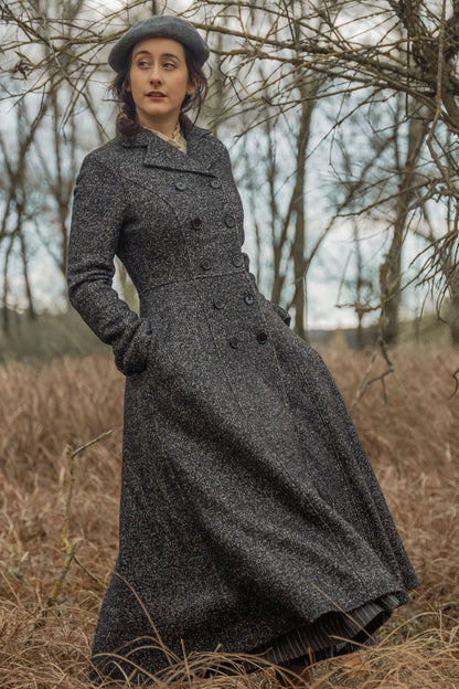 A woman wearing a long gray double-breasted wool maxi coat with notched collar, standing in a natural outdoor setting with trees in the background.
