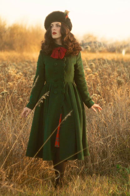 A woman standing in a field during sunset wearing a green wool winter dress coat with long sleeves, a lapel collar, and red accents.