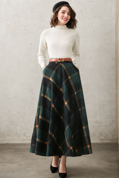 A woman standing in a studio setting, wearing a long winter wool plaid skirt in green, with a beige belt and a white sweater.