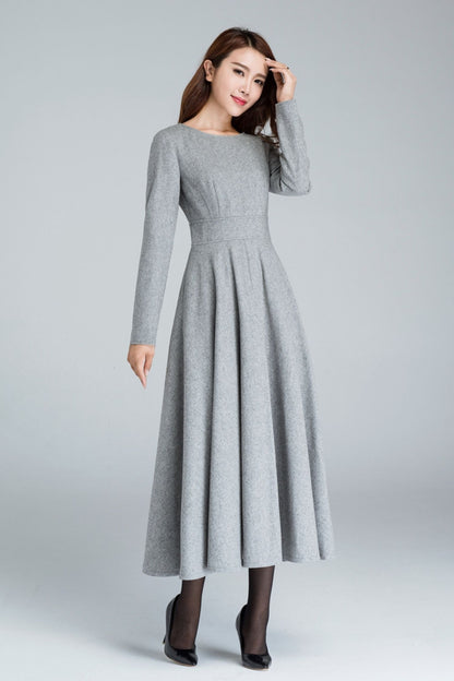 A woman standing, wearing a long sleeve gray wool swing dress with a round neckline and long skirt length.