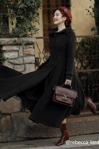 A woman wearing a long black wool swing coat with a hood, princess-style, with button fastening through the front, and two side seam pockets. The coat is mid-calf length, and the woman is posing on a stone bench with a building and tree in the background.