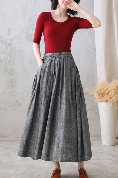 A woman wearing a red top and a gray pleated maxi linen skirt with pockets, standing in a room with a white chair and a vase in the background.