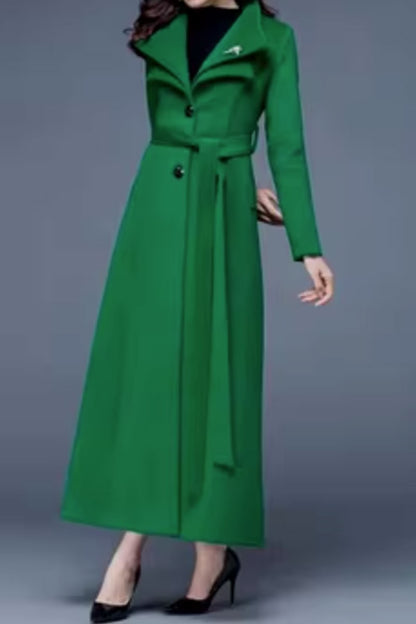 A woman standing wearing a green maxi winter wool warm coat with a belt waist and long sleeves. She has her hand in a pocket and is facing the camera.