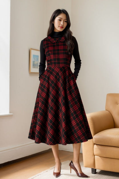A woman standing in a room wearing a sleeveless, V-neck, midi-length plaid wool dress in red and black, with a fitted waist and midi calf length.