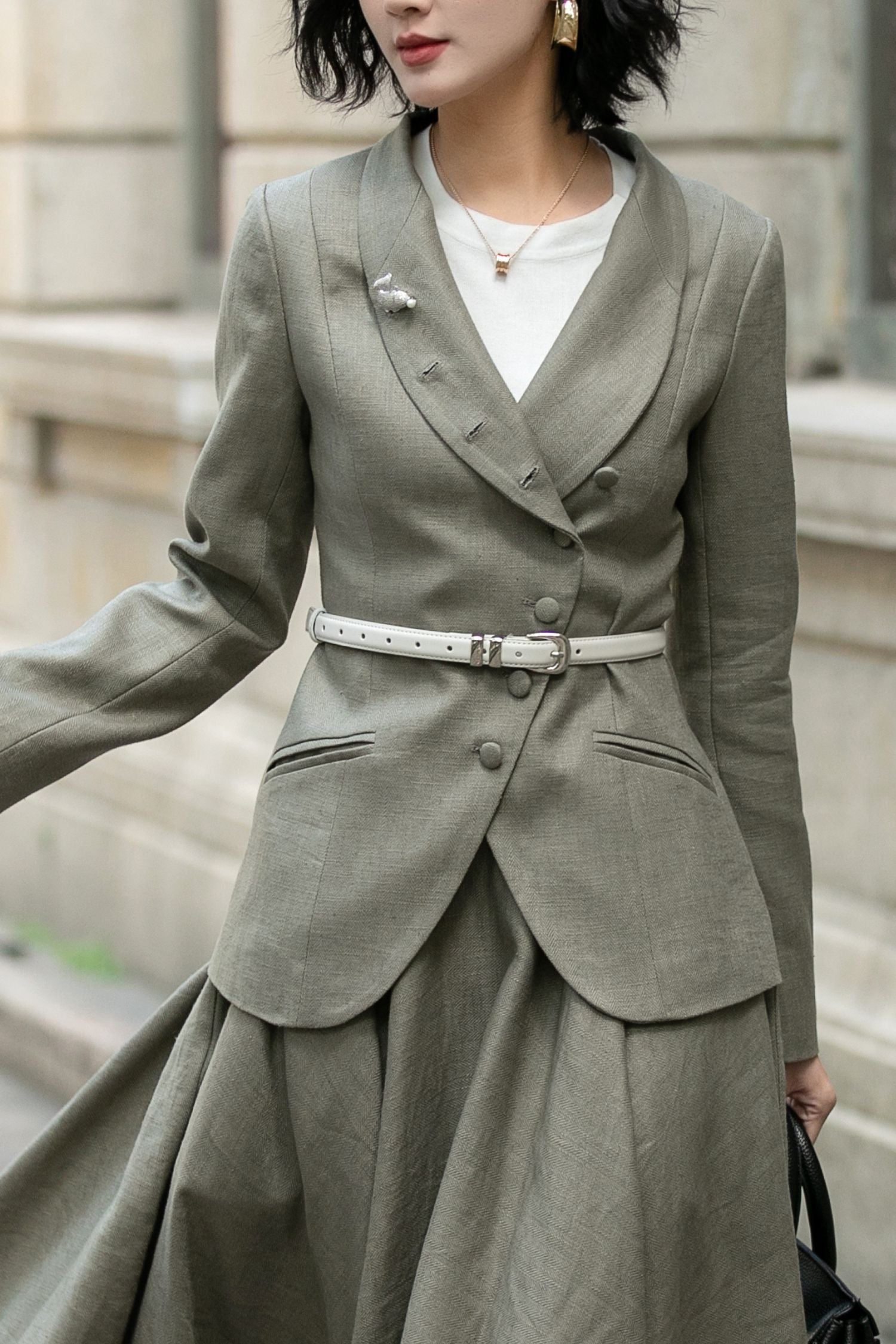 A woman wearing an olive green asymmetrical linen jacket with a button closure and outer lapel, paired with a light-colored top and a high-waisted midi skirt with pockets.