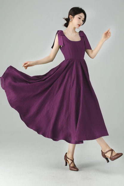 May include: A flowing, plum-colored dress with a square neckline and short sleeves. The dress features bow details on the shoulders and a flared skirt. The model is wearing brown heels.