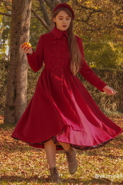 A woman wearing a red double-breasted wool princess coat with a cinched waist and full skirt, paired with an orange apple, walking in a park with autumn leaves on the ground.