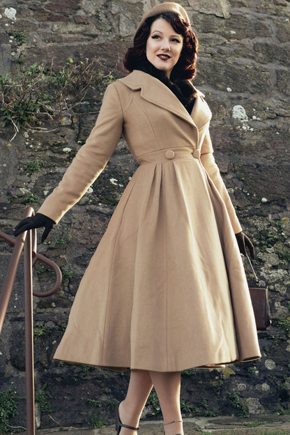 A woman wearing a vintage inspired beige wool Princess swing coat with long sleeves, a double-breasted button closure, and a pleated waist detail, standing on a pathway with walls in the background.