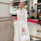 Woman in a white coat holding a white handbag on a city street.