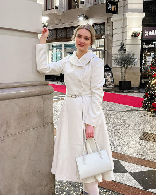 Woman in a white coat holding a white handbag on a city street.