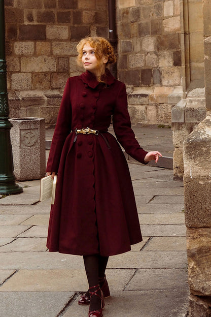 A woman wearing a burgundy double-breasted winter wool coat with a cinched waist and full skirt, standing on a street with historical buildings in the background.