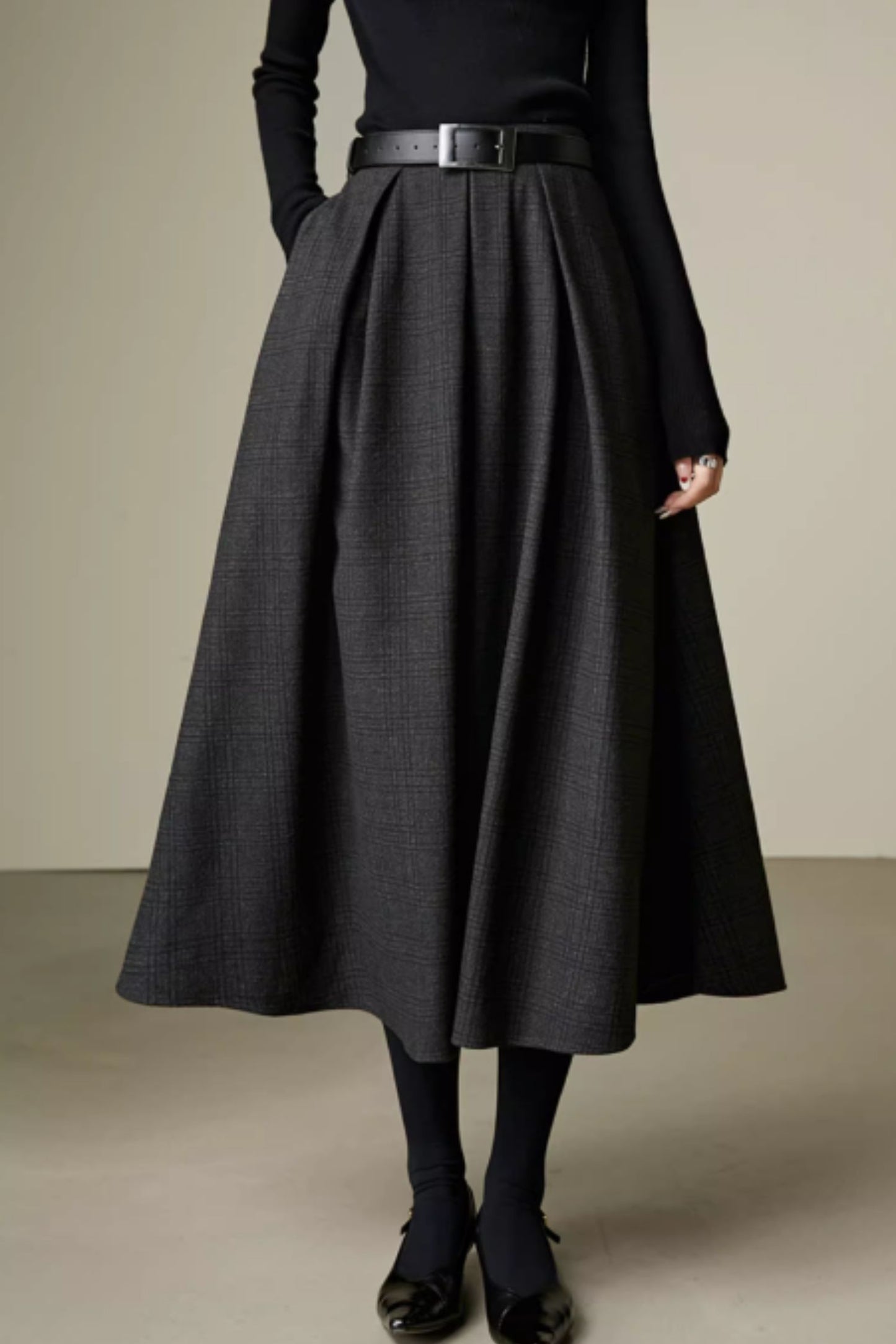 Person wearing a long dark gray skirt with a black top and shoes on a neutral background