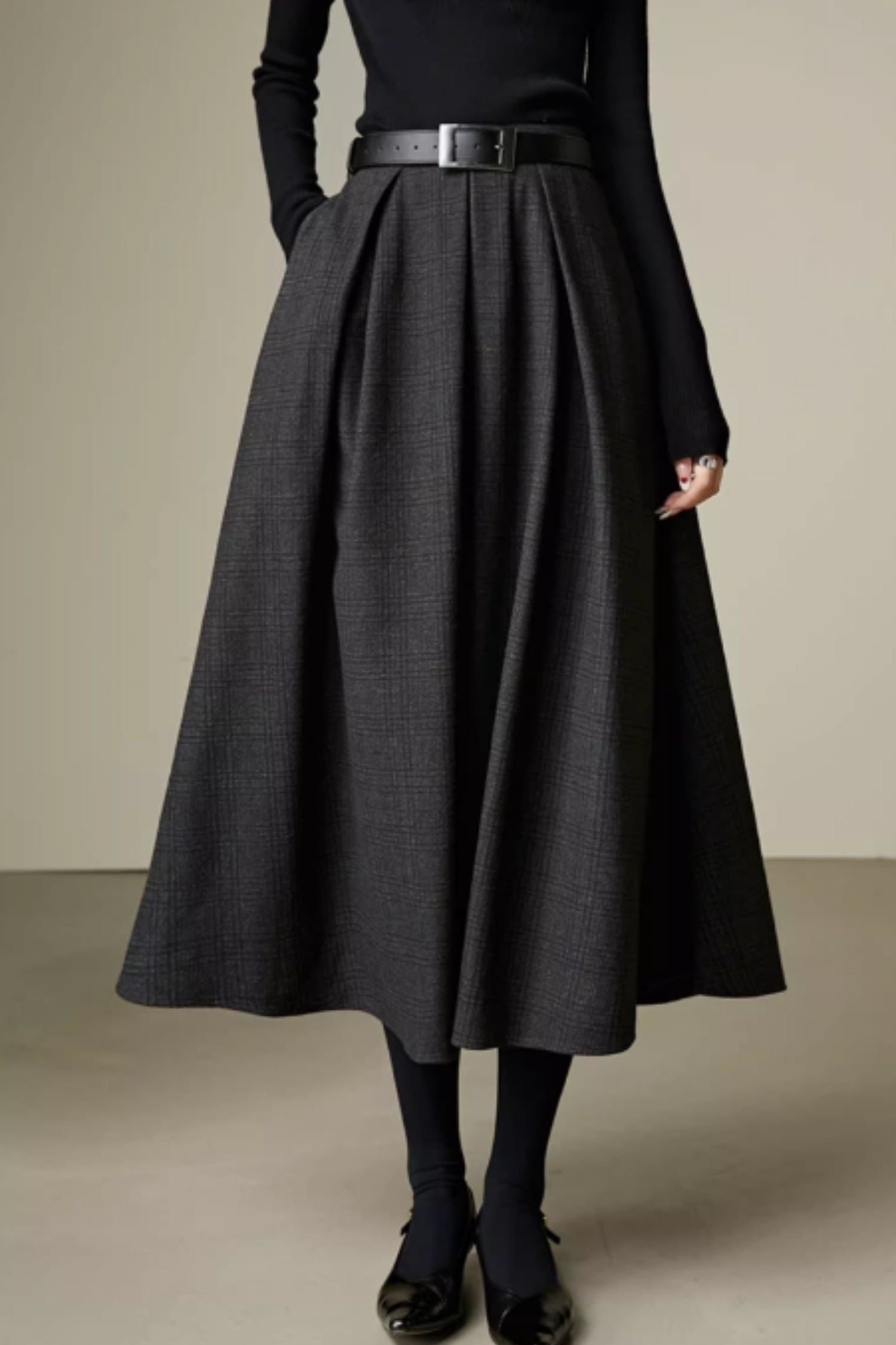 Person wearing a long dark gray skirt with a black top and shoes on a neutral background