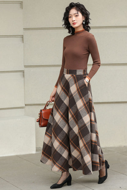 Woman wearing a brown turtleneck and plaid skirt holding a red handbag against a light-colored wall.