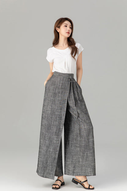 A model wearing gray wide leg asymmetrical linen wrap pants with a white top, standing against a neutral background.