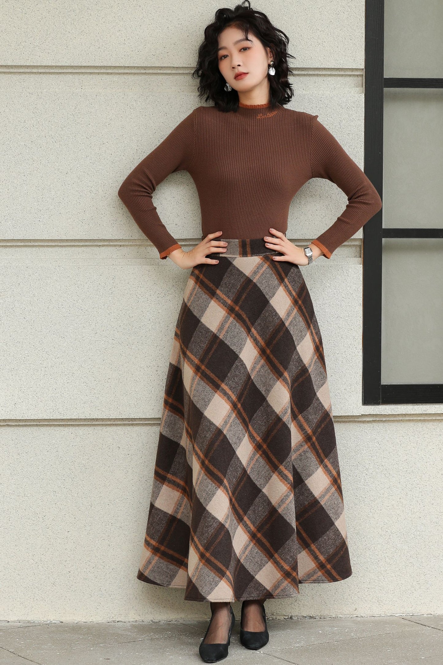Woman wearing a brown top and plaid skirt against a light-colored wall.
