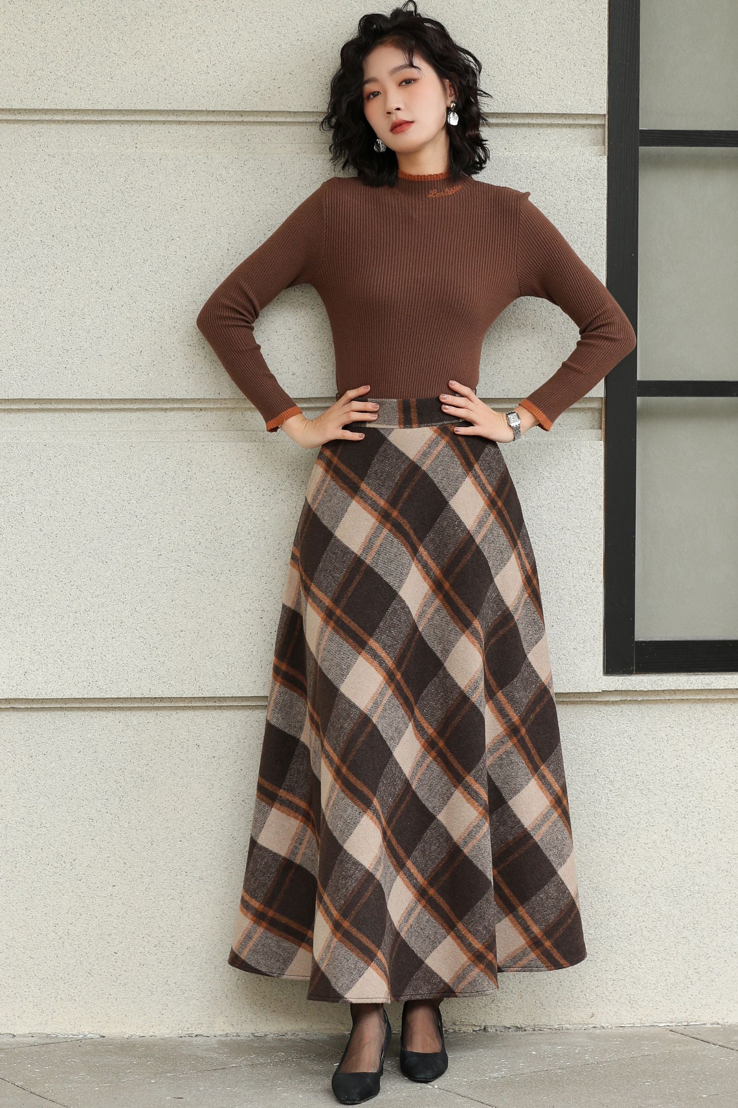 Woman wearing a brown top and plaid skirt against a light-colored wall.