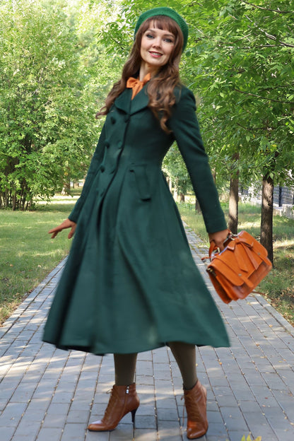 A woman standing outdoors wearing a green retro style wool coat with a calf-length skirt, brown leather shoes, and carrying an orange purse. The background is a tree-filled park.