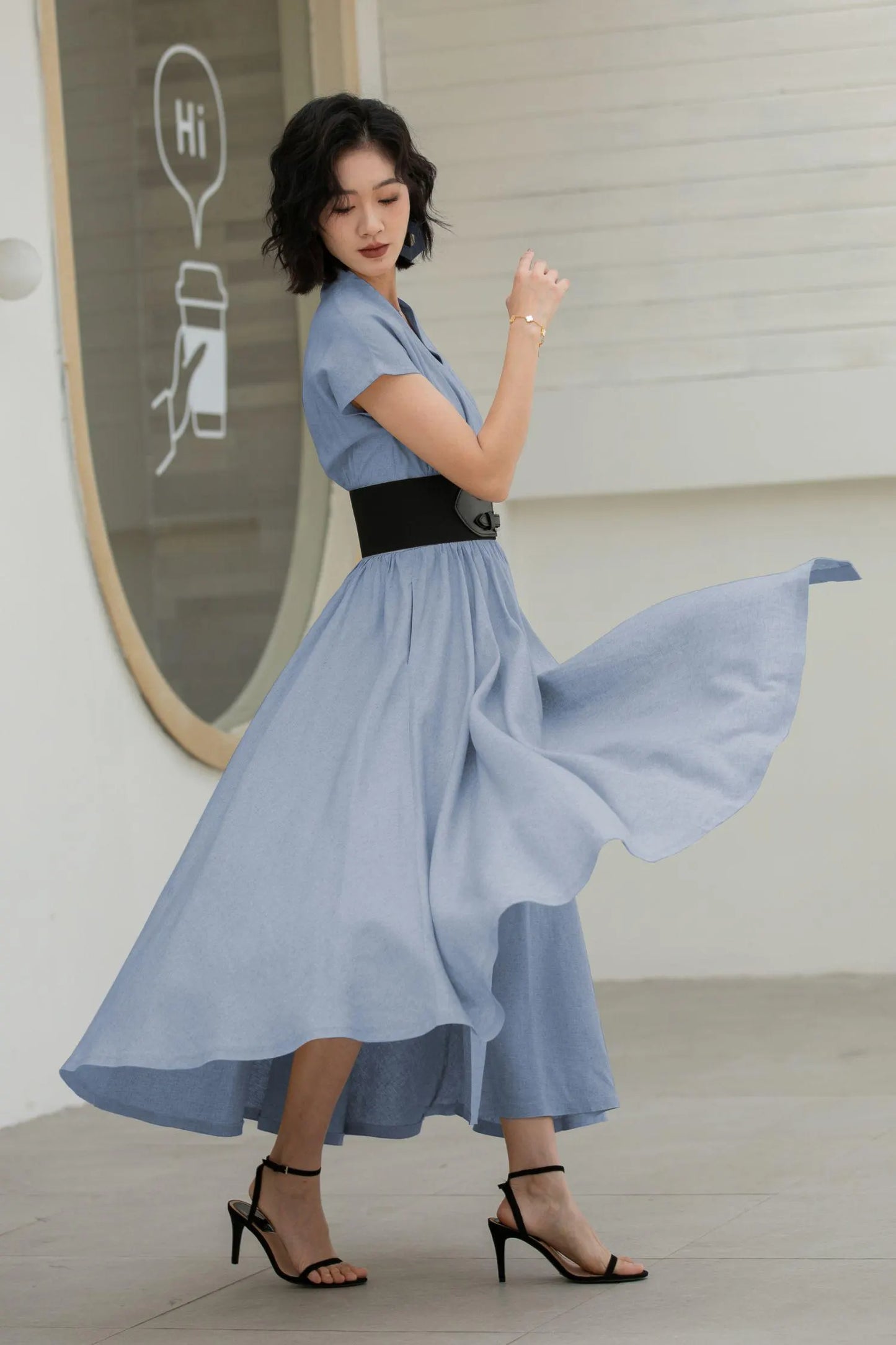 May include: A light blue dress with a V-neck and short sleeves, cinched at the waist with a wide black belt. The dress has a flowing skirt and is paired with black high-heeled sandals. The image also shows a white sign with the word "Hi".