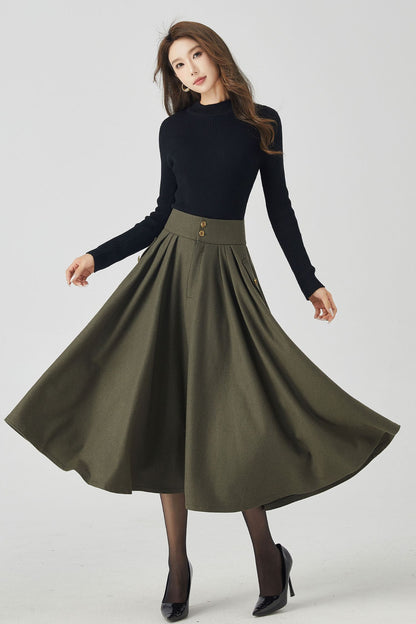 A woman modeling a military green pleated midi skirt with a black top and black boots.
