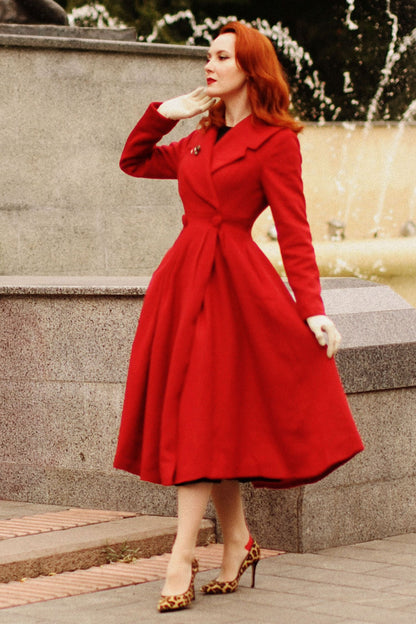 A woman standing outdoors wearing a red long princess wool coat with a lapel collar, button closure front, and mid calf length. She is also wearing high heels and a white glove.