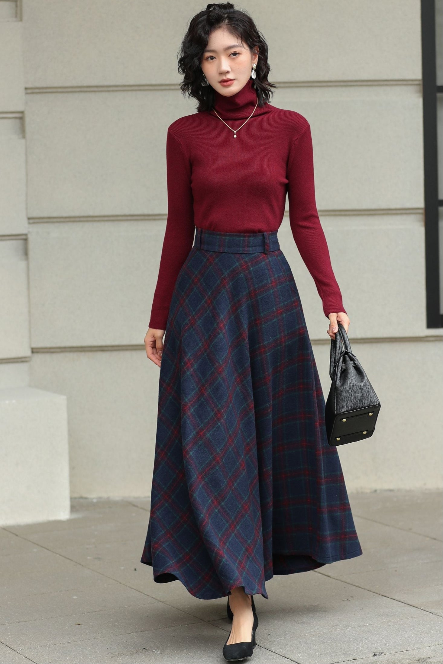 Woman wearing a red turtleneck and plaid skirt standing against a light-colored wall.