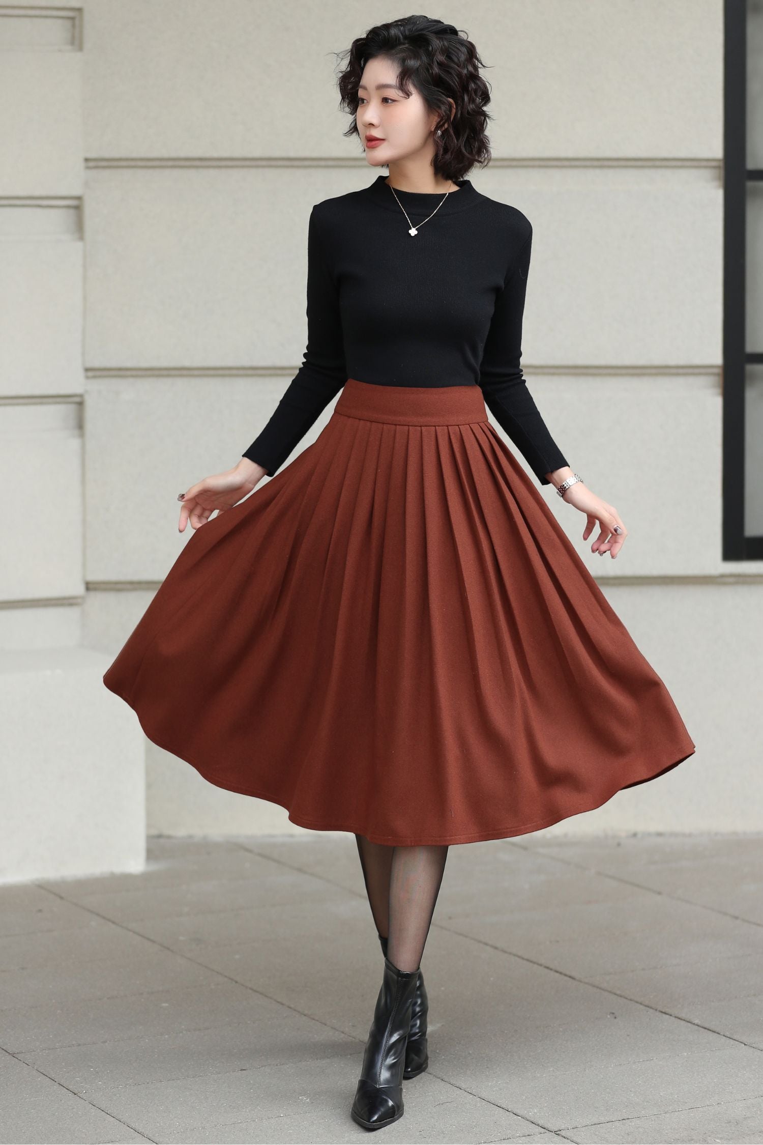 Woman wearing a black top and brown skirt standing against a light-colored wall.