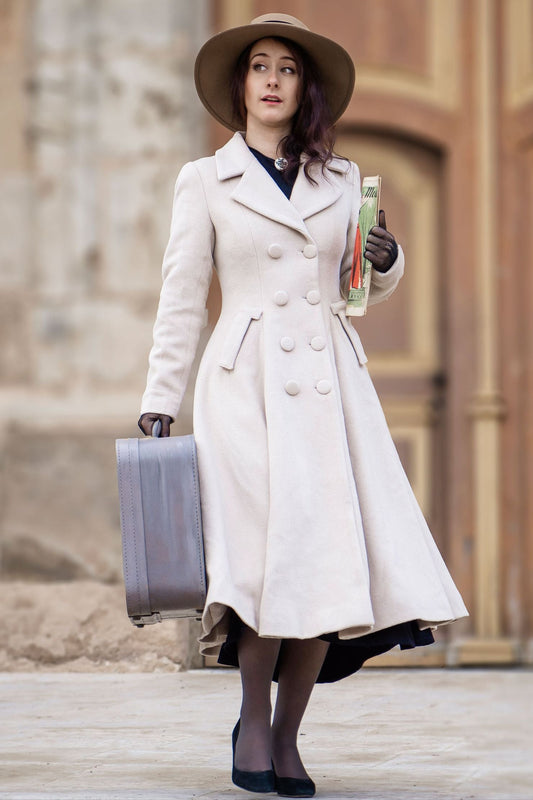 Woman in a long beige coat and hat holding a suitcase and a drink.