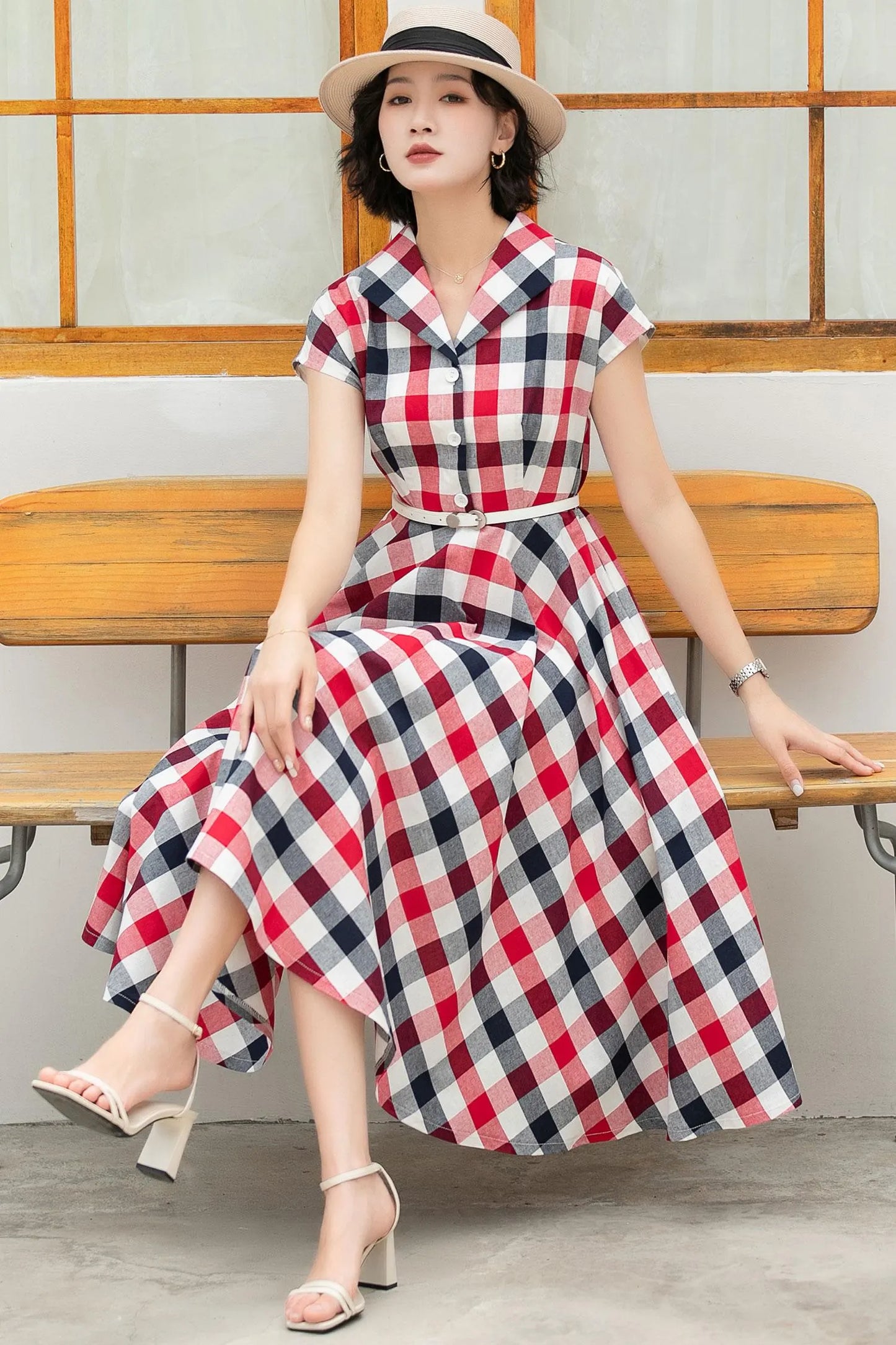 May include: A red, white, and blue plaid midi dress with short sleeves and a button-up front. The dress has a white belt and is worn with white heeled sandals.