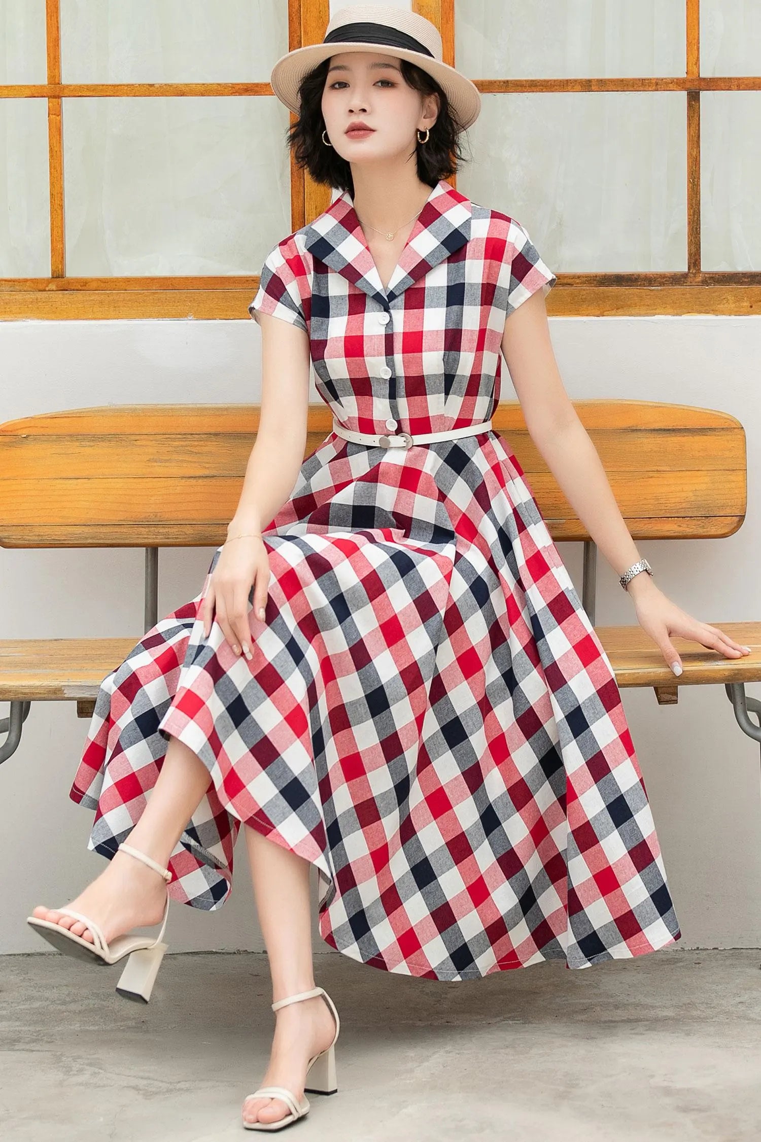 May include: A red, white, and blue plaid midi dress with short sleeves and a button-up front. The dress has a white belt and is worn with white heeled sandals.