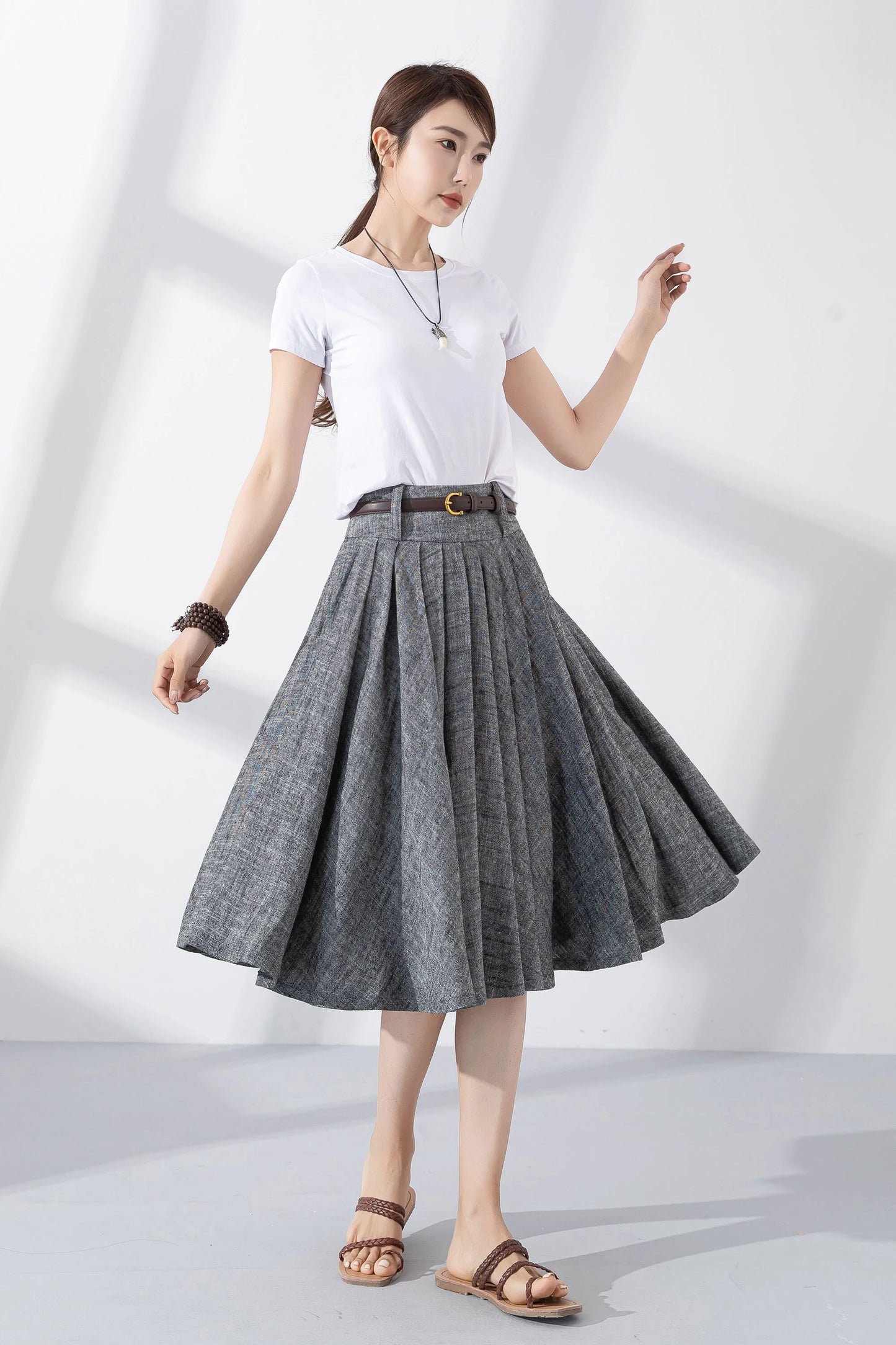 May include: A gray linen skirt with a brown belt and a gold buckle. The skirt is pleated and has a flared silhouette. The skirt is knee-length.
