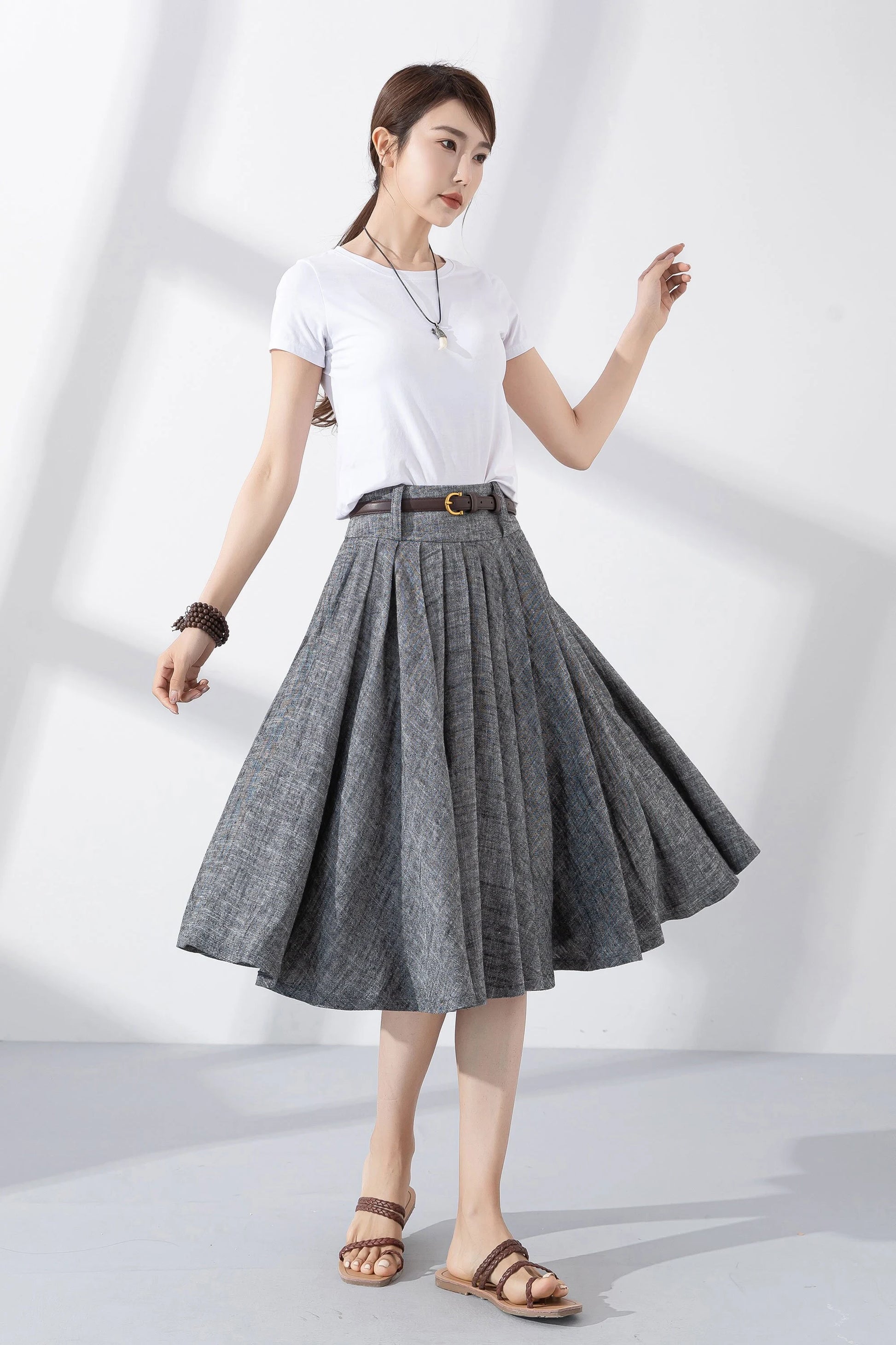 May include: A gray linen skirt with a brown belt and a gold buckle. The skirt is pleated and has a flared silhouette. The skirt is knee-length.