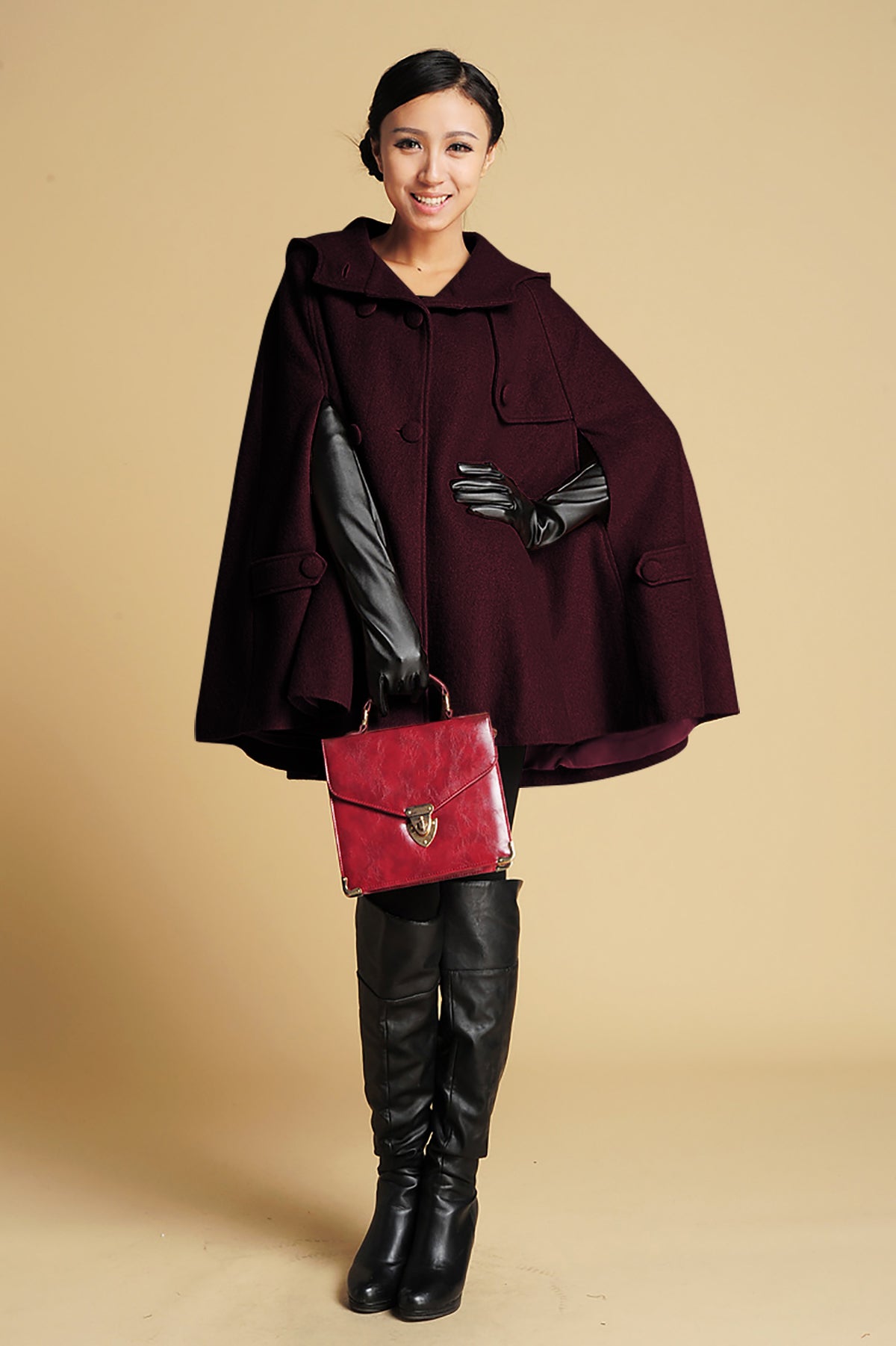Woman wearing a dark red cape with a black belt, holding a red handbag against a beige background