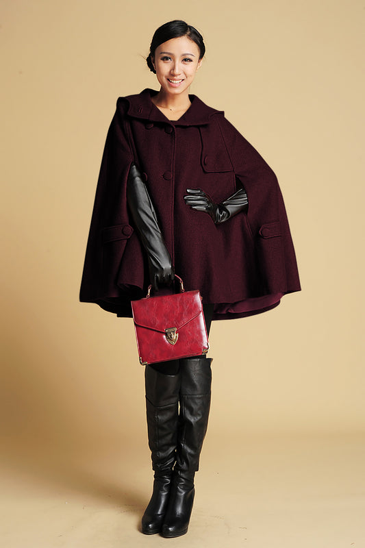 Woman wearing a dark red cape with a black belt, holding a red handbag against a beige background