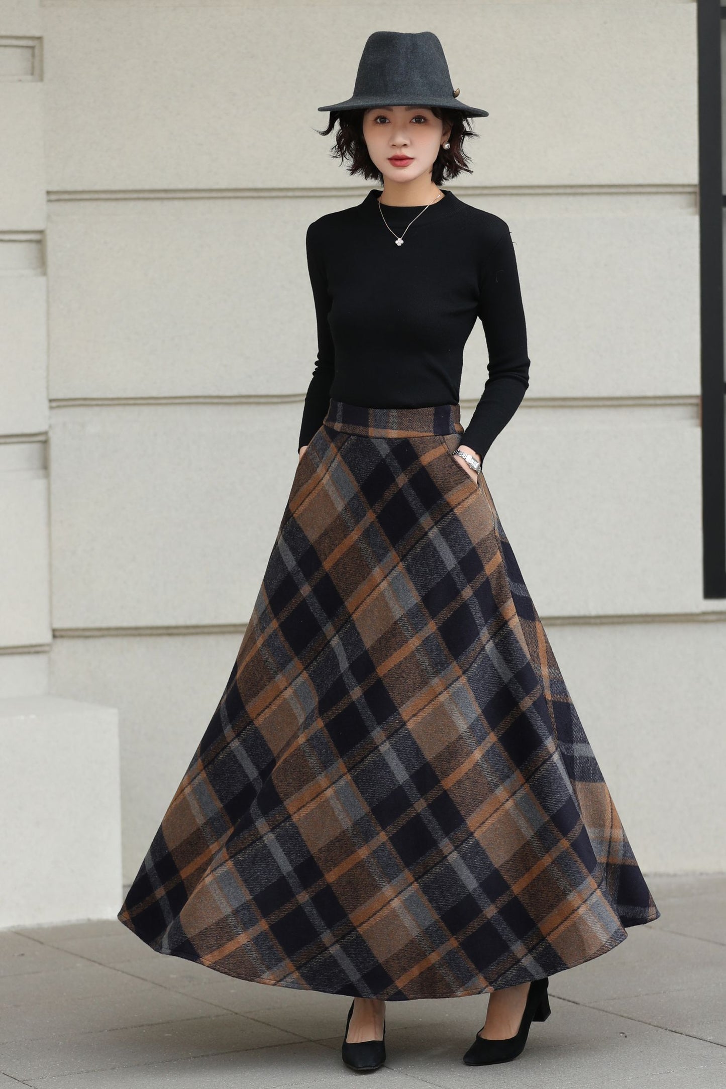 Woman wearing a black top and plaid skirt with a hat against a light-colored wall.