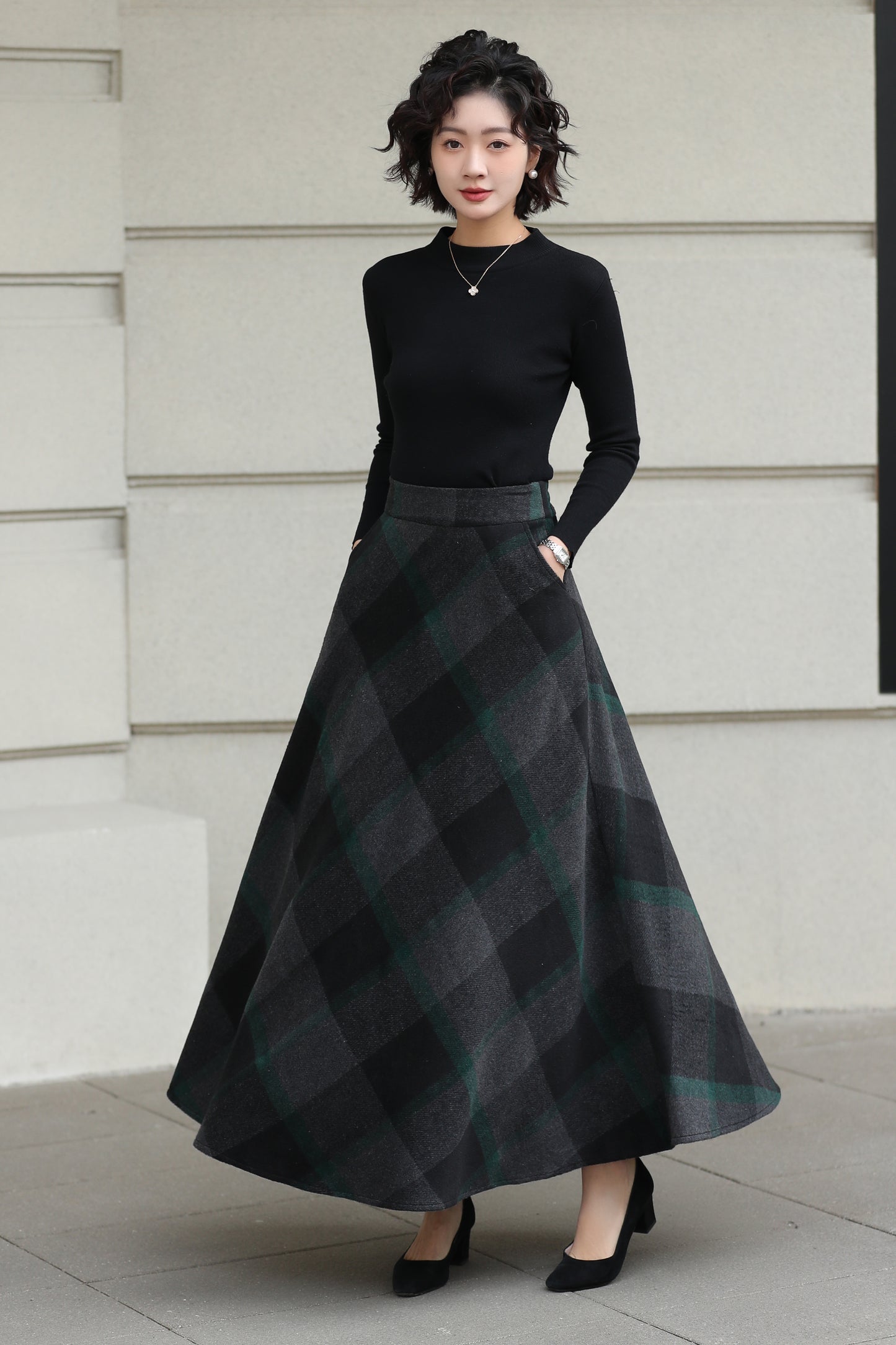 Woman wearing a black top and plaid skirt standing against a light-colored wall.