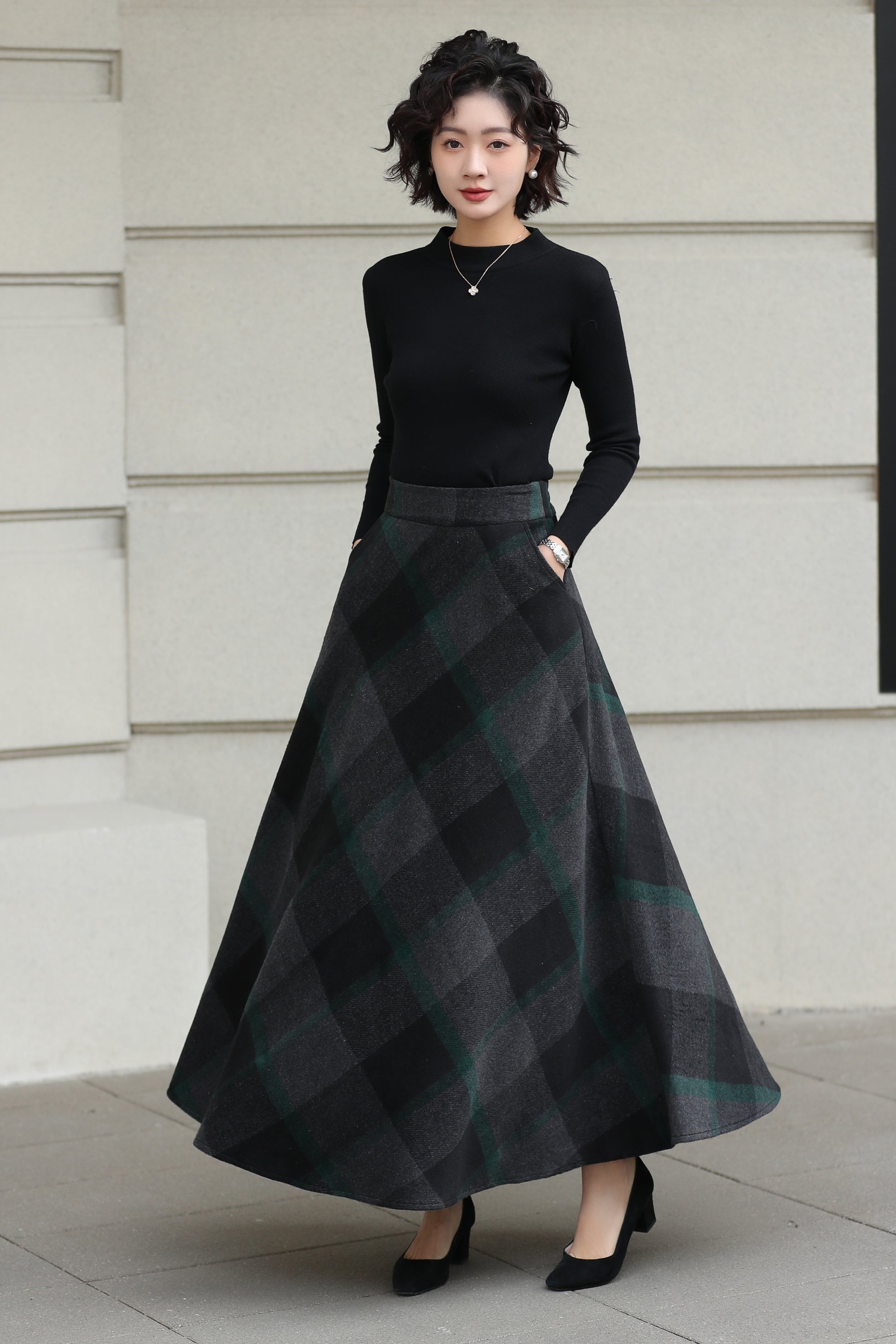 Woman wearing a black top and plaid skirt standing against a light-colored wall.