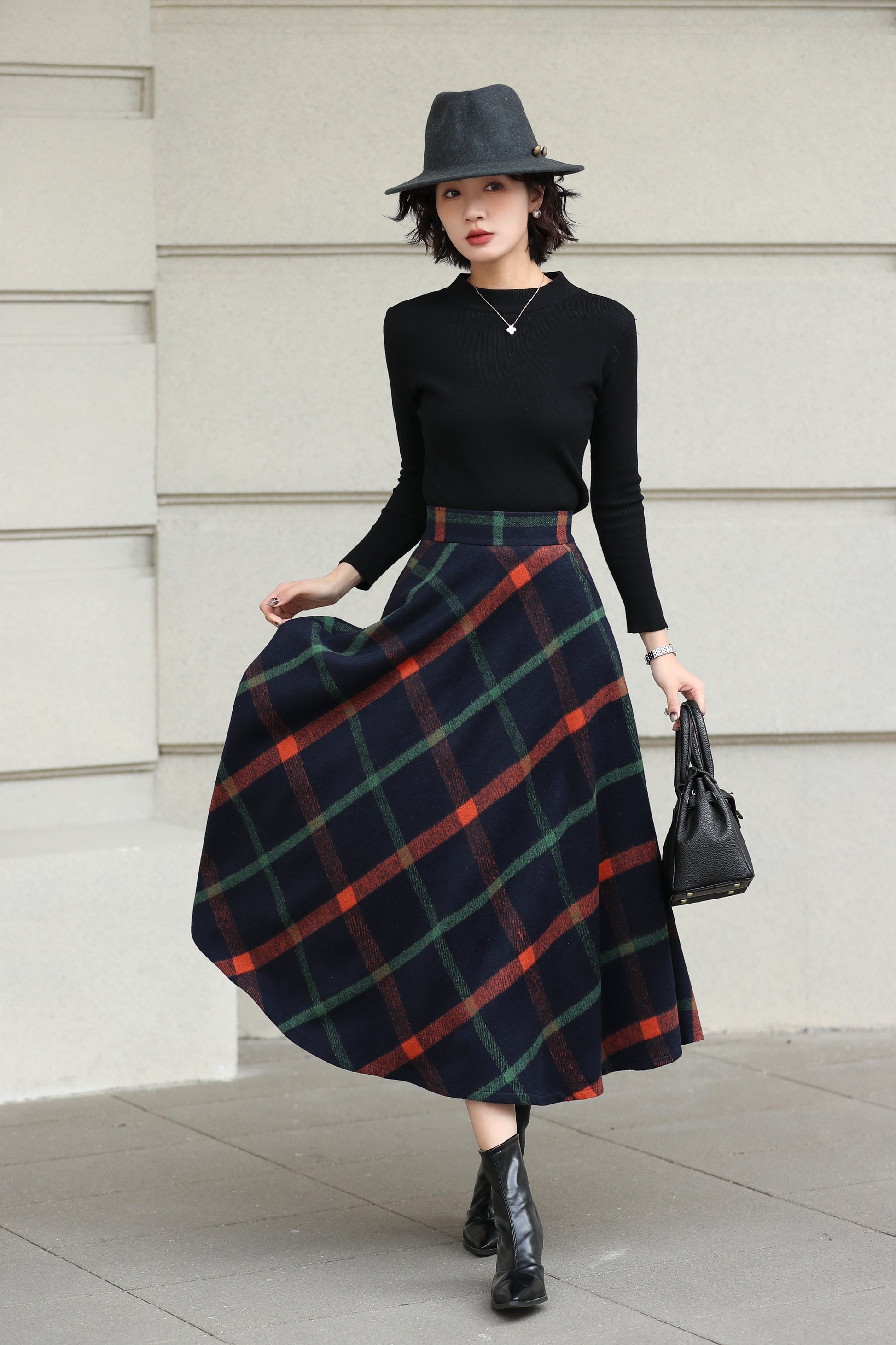 Woman wearing a black top, plaid skirt, and hat against a light-colored wall.