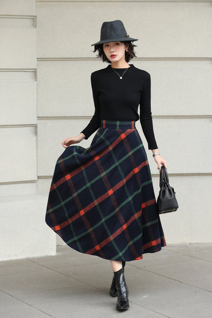 Woman wearing a black top, plaid skirt, and hat against a light-colored wall.