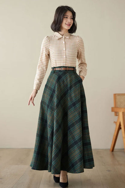 A woman wearing a green maxi wool plaid skirt with a checkered pattern, paired with a brown checkered coat and black shoes, standing in a room with a wooden floor and a chair in the background.